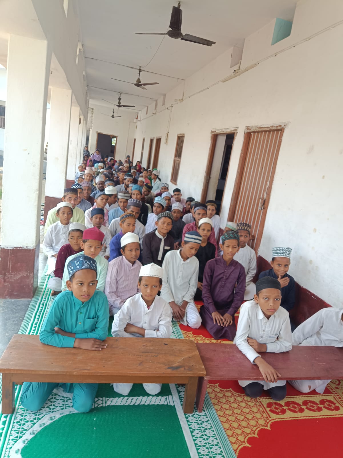 Students of Madarsa Gausia Badrul Uloom seated in rows on prayer mats in the veranda, facing the camera