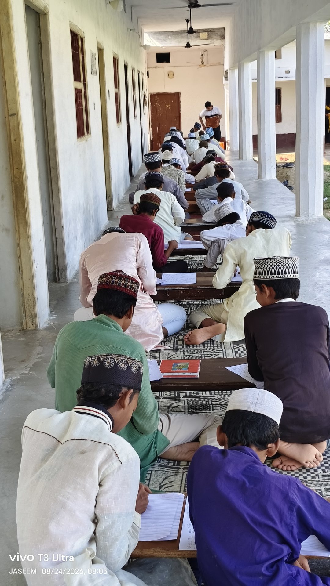 Students writing and studying at low desks in the veranda of the madarsa, seen from behind